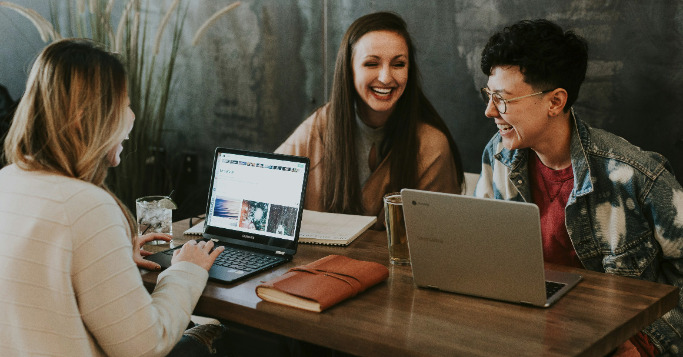 Drei Frauen sitzen an einem Tisch vor ihren Laptops und lachen gemeinsam.